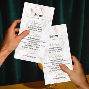 Könnte beinhalten: Zwei Menüs mit Marmoreffekt werden hochgehalten und zeigen die Angebote eines Restaurants. Die Menüs haben elegante goldene Schriftzüge und listen Vorspeisen, Hauptgerichte und Desserts auf. Der Hintergrund umfasst einen dunkelgrünen Vorhang und eine gelbe Oberfläche.