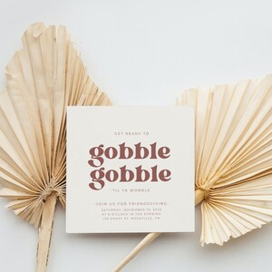 May include: A white card with brown text that reads "Get ready to gobble gobble 'til ya wobble." The card also includes the text "Join us for Friendsgiving - Saturday, November 19, 2022 at 8 o'clock in the evening 123 Cravy St. Nashville, TN". The card is sitting on top of two beige palm leaf fans.