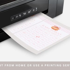 May include: A black printer printing a baby bingo game sheet. The sheet has a pink and white gingham border and features a pumpkin graphic. The text "BABY BINGO" is visible, along with other text in pink boxes. The bottom of the image reads "-PRINT FROM HOME OR USE A PRINTING SERVICE-".