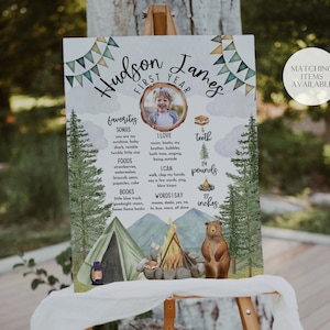 May include: A watercolor-style first birthday sign for a boy named Hudson James. The sign features a photo of the child, a campfire, a tent, a bear, and a list of his favorite things, including songs, foods, books, and words he says. The sign also includes his height and weight.