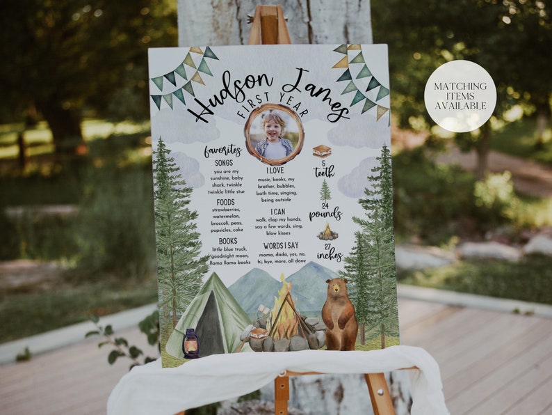 May include: A watercolor-style first birthday sign for a boy named Hudson James. The sign features a photo of the child, a campfire, a tent, a bear, and a list of his favorite things, including songs, foods, books, and words he says. The sign also includes his height and weight.
