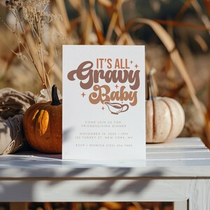 Op de afbeelding: Een witte kaart met bruine tekst die "It's All Gravy Baby" zegt met een juslepel-icoon. De kaart is een uitnodiging voor een Friendsgiving-diner met de datum, tijd en locatie erop gedrukt. De kaart ligt op een witte tafel met twee pompoenen op de achtergrond.