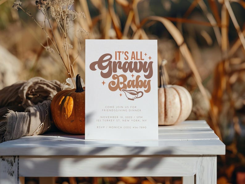 Op de afbeelding: Een witte kaart met bruine tekst die "It's All Gravy Baby" zegt met een juslepel-icoon. De kaart is een uitnodiging voor een Friendsgiving-diner met de datum, tijd en locatie erop gedrukt. De kaart ligt op een witte tafel met twee pompoenen op de achtergrond.