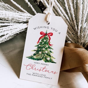 May include: A white gift tag with a watercolor Christmas tree design. The tag reads "Wishing You a Very Merry Christmas" in red script. A red bow tops the tree. The tag is tied with a white string.