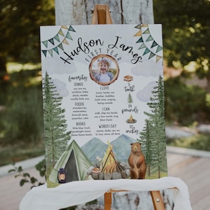 May include: A watercolour-style poster with a woodland theme celebrating a child's first birthday. The poster features a photo of the child, a list of their favourite songs, foods, and books, and their height and weight. The poster is decorated with a campfire, a tent, and a bear.