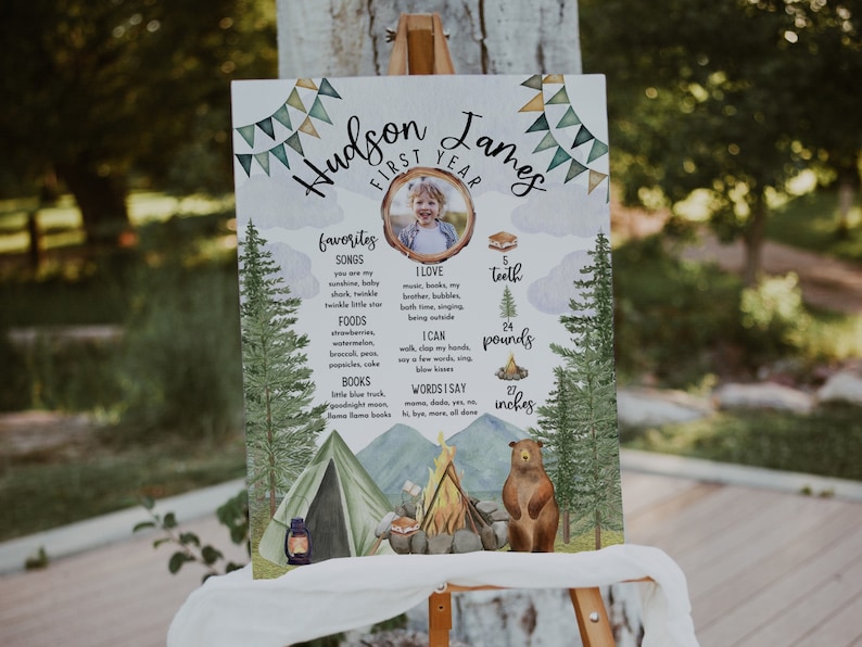 May include: A watercolour-style poster with a woodland theme celebrating a child's first birthday. The poster features a photo of the child, a list of their favourite songs, foods, and books, and their height and weight. The poster is decorated with a campfire, a tent, and a bear.