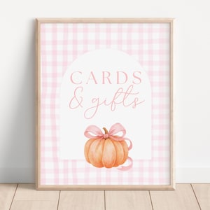 May include: A framed sign with a pink gingham background. The sign features the words "CARDS & gifts" in pink script within a white arch. Below the text is an orange pumpkin with a pink bow and ribbon.