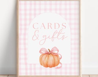 May include: A framed sign with a pink gingham background. The sign features the words "CARDS & gifts" in pink script within a white arch. Below the text is an orange pumpkin with a pink bow and ribbon.
