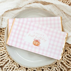May include: Two chocolate bars with pink gingham wrappers and gold edges rest on a gray plate. Each wrapper features a label with the text "LAYLA'S first birthday" and a pumpkin illustration.