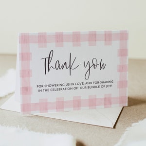May include: A white thank you card with a pink and white gingham pattern border. The text on the card reads "Thank you for showering us in love, and for sharing in the celebration of our bundle of joy!"
