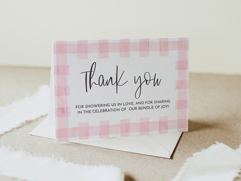 May include: A white thank you card with a pink and white gingham pattern border. The text on the card reads "Thank you for showering us in love, and for sharing in the celebration of our bundle of joy!"