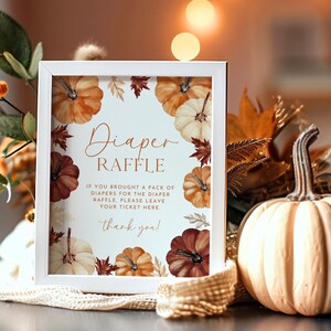 May include: A white framed sign with the words "Diaper Raffle" surrounded by watercolor pumpkins and fall leaves. A decorative pumpkin and fall foliage are in the background. The sign reads "If you brought a pack of diapers for the diaper raffle, please leave your ticket here. Thank you!"