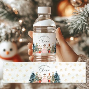 Könnte beinhalten: Wasserflaschenetikett mit Nussknacker-Motiv. Das Etikett zeigt eine Aquarell-Illustration von Clara und dem Nussknacker vor einem Weihnachtsbaum. Der Text auf dem Etikett lautet "Happy Birthday Clara".