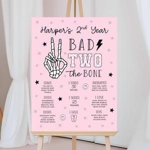 Puede incluir: Cartel de cumpleaños rosa con el texto "Harper's 2nd Year" y "Bad Two the Bone". El cartel incluye una mano de esqueleto haciendo el signo de la paz, estrellas y rayos, y detalles sobre la cumpleañera, incluyendo su altura de 76 cm y su peso de 10 kg.
