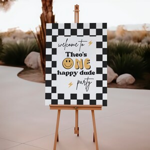 Puede incluir: Un cartel blanco con fondo de cuadros negros y blancos. El cartel dice "Bienvenido a la fiesta de Theo's One happy dude" con una carita sonriente amarilla y rayos.