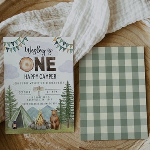 May include: A birthday party invitation for a one-year-old boy with a woodland camping theme. The invitation features a brown bear, a campfire, a tent, and a sign that says "Wesley is One Happy Camper". The invitation also includes the date, time, and location of the party.