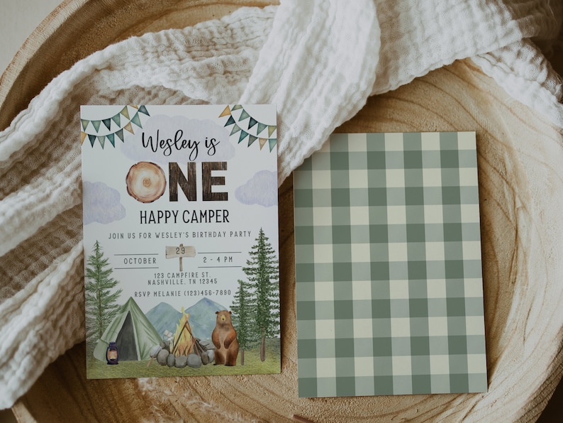 May include: A birthday party invitation for a one-year-old boy with a woodland camping theme. The invitation features a brown bear, a campfire, a tent, and a sign that says "Wesley is One Happy Camper". The invitation also includes the date, time, and location of the party.
