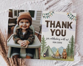 May include: A baby in a brown beanie and plaid shirt smiles in a high chair next to a thank you card. The card features a camping scene with a tent, campfire, bear, and the words "THANK YOU for celebrating with us!"