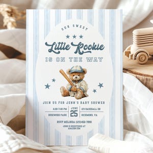 May include: Baby shower invitation with a baseball theme. The invitation features a teddy bear wearing a baseball uniform and holding a bat. The text reads "Our Sweet Little Rookie is on the Way". The background has blue and white stripes.