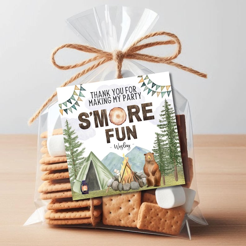 May include: A clear plastic bag filled with graham crackers, marshmallows, and chocolate squares. A white tag with the text "Thank you for making my party S'more Fun - Wesley" is attached to the bag. The tag features a watercolor illustration of a campfire, tent, and trees.