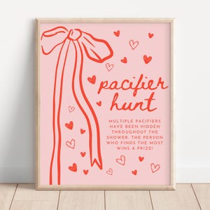 Może przedstawiać: Różowy znak gry na baby shower z czerwonym tekstem i ilustracjami. Znak głosi "pacifier hunt" ze wstążką i sercami. Tekst poniżej wyjaśnia zasady gry. Znak jest oprawiony w jasną drewnianą ramę.