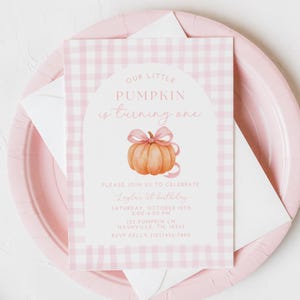 May include: A pink and white gingham-patterned invitation card with the text "Our Little Pumpkin is turning one." The card features a watercolor pumpkin with a pink bow. The invitation is on a pink plate with a white envelope.
