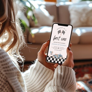 May include: A smartphone displaying a birthday invitation for a first birthday party. The invitation features a blue race car with a checkered flag background and the text "Race on over to celebrate Hudson's 1st Birthday".
