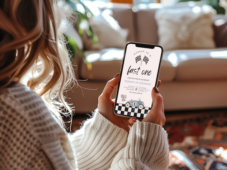 May include: A smartphone displaying a birthday invitation for a first birthday party. The invitation features a blue race car with a checkered flag background and the text "Race on over to celebrate Hudson's 1st Birthday".
