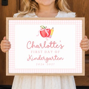 May include: A rectangular wooden frame containing a sign with a light pink gingham border. The center features a watercolor red apple with a pink ribbon and a yellow pencil, with the text Charlotte's FIRST DAY OF Kindergarten 2026-2027.