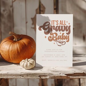 Op de afbeelding: Een witte kaart met de tekst "It's All Gravy Baby" in bruine en oranje letters. De kaart ligt op een houten tafel met twee pompoenen en een kleine kalebas.