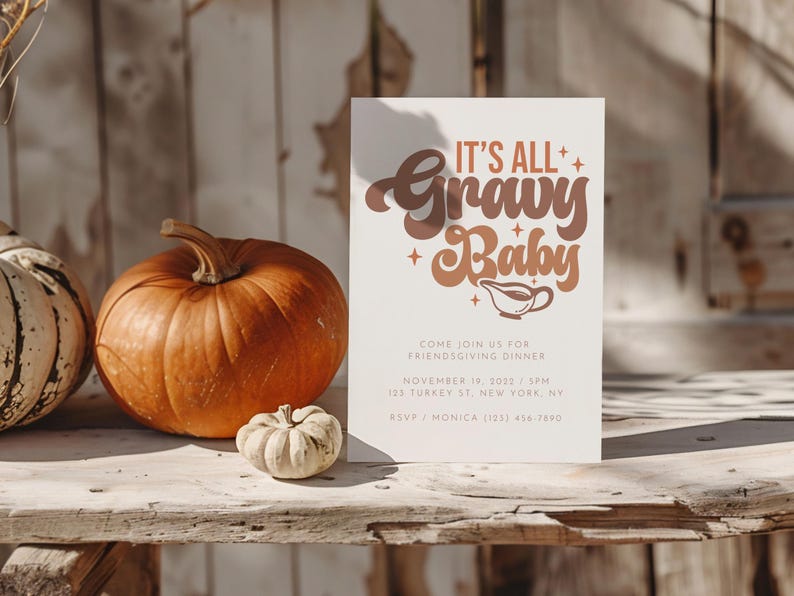 Op de afbeelding: Een witte kaart met de tekst "It's All Gravy Baby" in bruine en oranje letters. De kaart ligt op een houten tafel met twee pompoenen en een kleine kalebas.