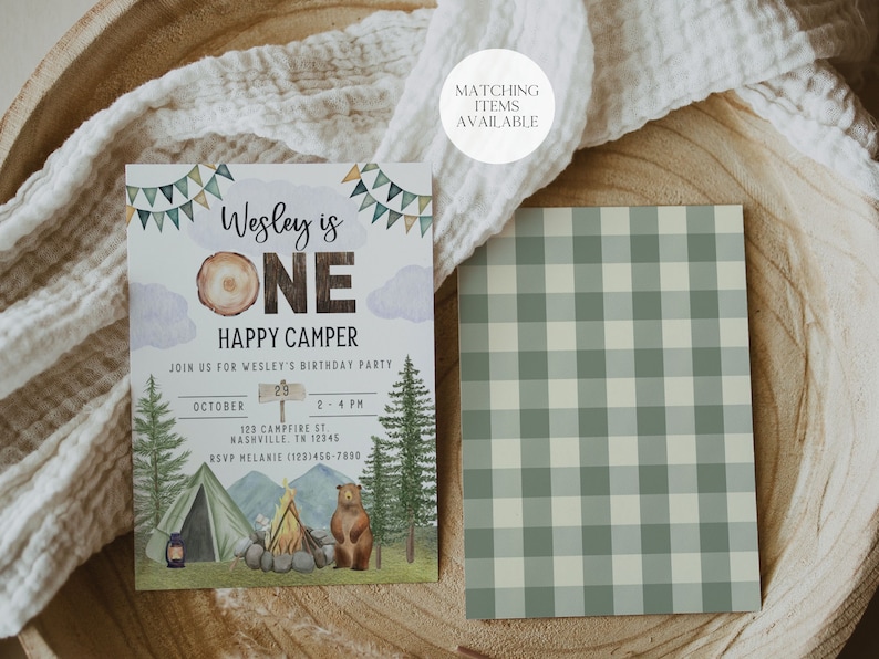May include: A green and white gingham pattern with a birthday invitation for a boy's first birthday party. The invitation features a woodland theme with a bear, a campfire, a tent, and a sign that says "Wesley is One Happy Camper". The invitation also includes the date, time, and location of the party.