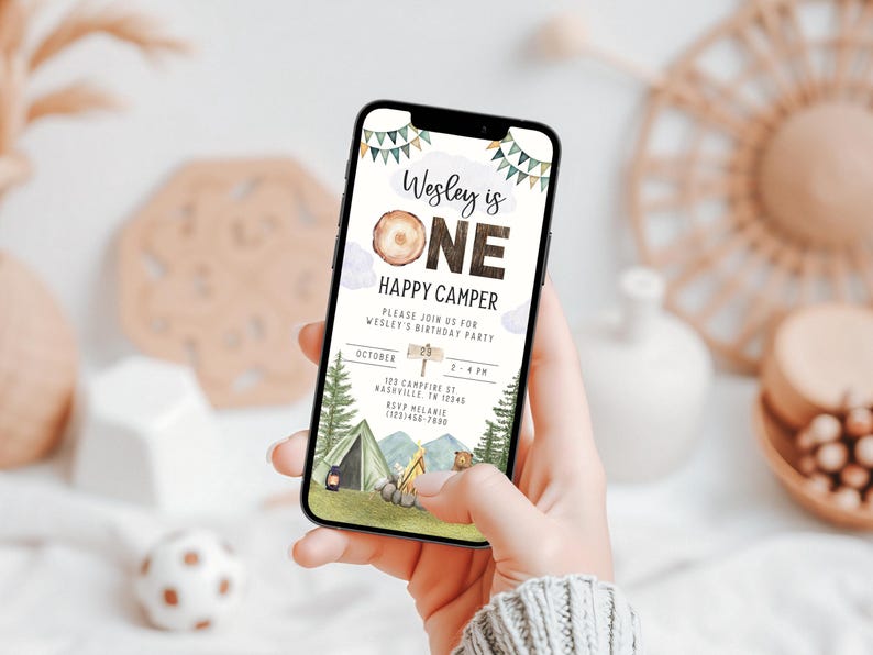 May include: A digital invitation for a first birthday party with a woodland camping theme. The invitation features a wood-grain background with the text "Wesley is ONE" and "Happy Camper". The invitation also includes details about the party, such as the date, time, and location.