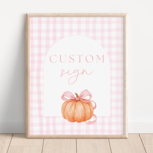 May include: Framed print with a watercolour pumpkin and pink bow on a pink gingham backdrop. The words "CUSTOM sign" are in pink within a white arch. The design evokes autumn and fall themes, suitable for seasonal home decor.
