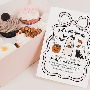 May include: A pink box of cupcakes and a Halloween-themed invitation. The invitation reads "Let's get spooky" and includes a ghost, pumpkin, and black cat illustration. Cupcakes have various toppings.