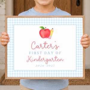 May include: A rectangular wooden frame containing a sign with a light blue gingham border. The sign features a watercolor red apple and yellow pencil, with the text: Carter's FIRST DAY OF Kindergarten 2026-2027 in red and blue lettering.