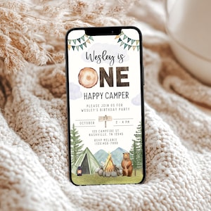 May include: A digital invitation for a first birthday party with a woodland camping theme. The invitation features a brown bear, a campfire, a tent, and a sign that says "Wesley is One Happy Camper". The invitation also includes the date, time, and location of the party.