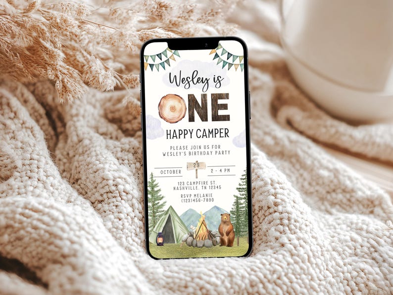 May include: A digital invitation for a first birthday party with a woodland camping theme. The invitation features a brown bear, a campfire, a tent, and a sign that says "Wesley is One Happy Camper". The invitation also includes the date, time, and location of the party.