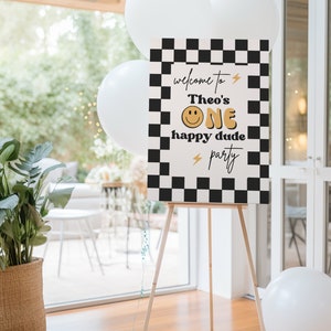 Puede incluir: Un cartel de bienvenida a cuadros blanco y negro con el texto "Bienvenido a la fiesta de Theo's One happy dude" en letras doradas y blancas. Se incluye una carita sonriente en el texto.