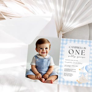 May include: A baby boy in a blue romper smiles next to a birthday invitation. The invitation has a blue gingham border and features a cartoon goose holding a balloon. The text reads "Campbell is One silly goose."