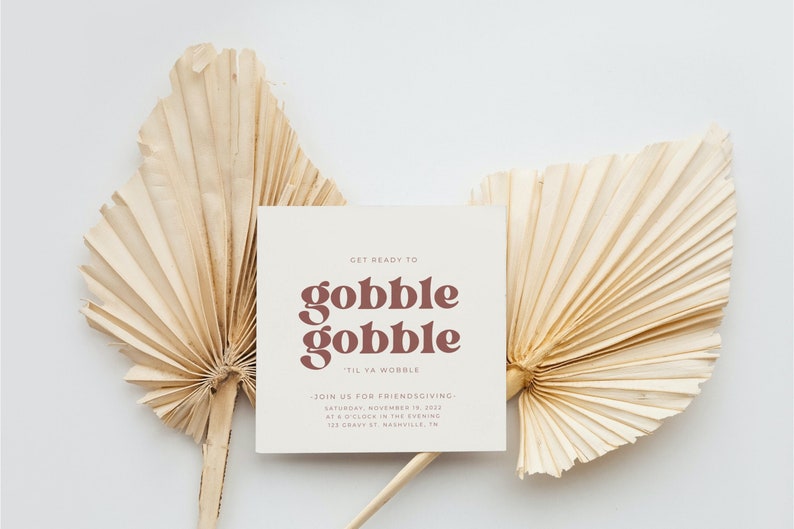 May include: A white card with brown text that reads "Get ready to gobble gobble 'til ya wobble." The card also includes the text "Join us for Friendsgiving - Saturday, November 19, 2022 at 8 o'clock in the evening 123 Cravy St. Nashville, TN". The card is sitting on top of two beige palm leaf fans.