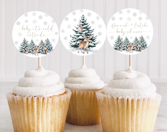 May include: Three white cupcake toppers with a winter woodland theme. The toppers feature a deer, a squirrel, and a bird in a snowy forest setting. The text on the toppers reads "Baby it's cold outside" and "Havah & Caleb's Baby Shower".
