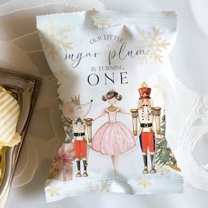 May include: White favor bag with a Nutcracker theme. It features a ballerina, a mouse king, and a Nutcracker soldier. The bag has gold snowflakes and the text "Our little sugar plum is turning ONE".