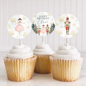May include: Three cupcake toppers featuring a Nutcracker theme. The first topper shows a ballerina in a pink tutu with snowflakes and gold glitter. The second topper shows a Nutcracker soldier with a Christmas tree and the text "Happy Birthday Clara". The third topper shows a Nutcracker soldier with snowflakes and gold glitter.
