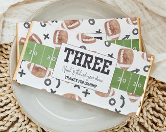 May include: Two chocolate bars with football-themed wrappers. The wrappers feature brown footballs, green yard lines, and the text "THREE Noah's third year down THANKS FOR COMING!" on a white background. The bars are on a plate.