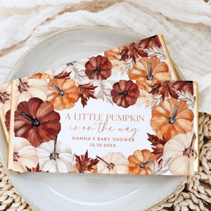 May include: Two chocolate bar wrappers with a fall-themed design. The wrappers feature a border of pumpkins in shades of orange, brown, and white, along with fall leaves. The text "A Little Pumpkin is on the Way" is displayed, along with baby shower details.