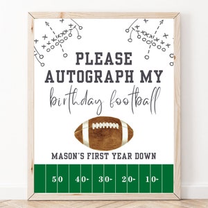 May include: A white sign with a football graphic and the text "PLEASE AUTOGRAPH MY birthday football" and "MASON'S FIRST YEAR DOWN". The sign has a green football field graphic with yard lines marked 50, 40, 30, 20, and 10.