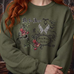 Peut inclure: Un sweat-shirt à col rond vert olive avec un imprimé graphique composé de roses rouges, d'ailes d'ange blanches et du texte By the Angel et All the Stories Are True. Le motif inclut des volutes noires ornées et divers symboles gothiques.