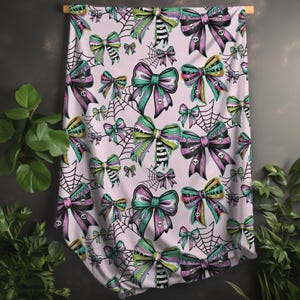 May include: A light pink fabric with a repeating pattern of colorful bows, spiderwebs, and skulls. The bows are in shades of green, purple, and yellow. The spiderwebs are black. The fabric is hanging from a wooden rod.