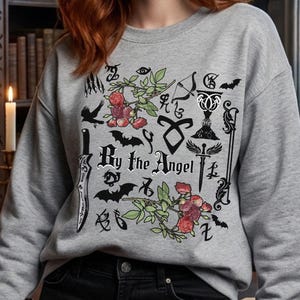 Peut inclure: Un sweat-shirt gris chiné à col rond avec une impression graphique noire portant le texte By the Angel. Le motif comprend des illustrations d'un couteau, de chauves-souris, de roses, d'un calice, d'une épée et divers symboles runiques.